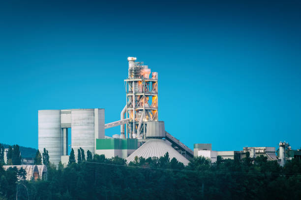 Horizontal composition cold colors photography of exterior of factory, industrial building at water's edge and at twilight time of the day, blue hour, with dark blue clear sky with copy space. This is a cement factory, concrete industry manufacturing. This picture was taken in Montalieu-Vercieu town, in Isere, in Auvergne-Rhone-Alpes region in France (Europe), along the Rhone river.